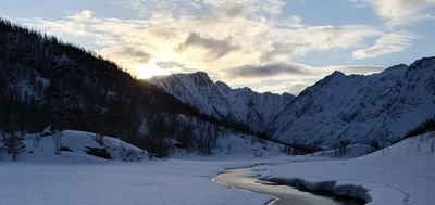 Snow-covered mountain landscape with a river, surrounded by high mountains and cloudy sky at sunset.