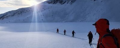 People in red jackets are hiking with ski poles through a snowy mountain landscape at sunrise.