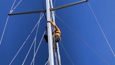 Person in yellow jacket climbing on a sailboat mast, secured with a climbing harness, against a blue sky.