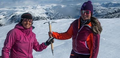 Two people in winter clothing with sunglasses and ice axes on a snow-covered mountain peak.