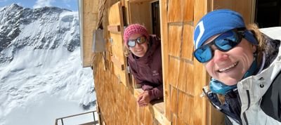 Two people with sunglasses are looking out of a wooden window at the snow-covered mountains.