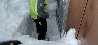 Person in a green jacket stands in the snow in front of a metal door, surrounded by high snow walls.