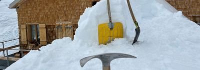 Ice pick and shovel are stuck in the snow in front of a mountain hut with a wooden facade.