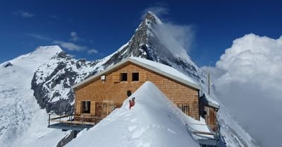 Wooden cabin on a snow-covered mountain peak with surrounding snowy mountains in the background.