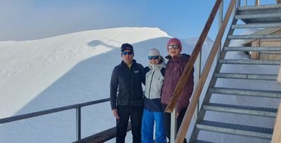 Three people in winter clothing with sunglasses are standing on a wooden platform in the snow.