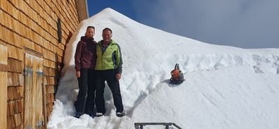 Two people are standing in winter clothing next to a snow-covered building in the mountains.