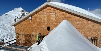 Person in front of a mountain hut in the snow, surrounded by snow-covered mountains, in sunny weather.