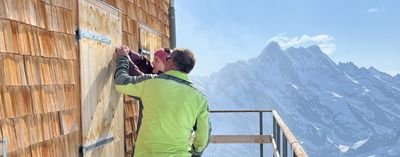 People in outdoor clothing are opening a wooden door at a mountain hut, with snow-covered mountains in the background.