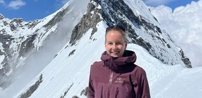 Person in purple jacket stands in front of snow-covered mountain in the Alps, blue sky in the background.