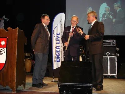 Three men in suits on a stage with microphones and a banner in the background.
