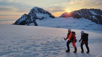 Two people with helmets and crampons are hiking in the snow in front of a mountain backdrop at sunrise.
