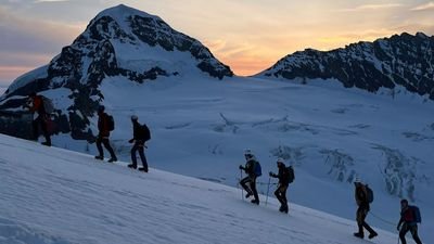 Group of people with helmets and sticks ascending a snow-covered mountain at sunset.