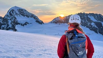 Person with helmet and backpack standing on a snow-covered mountain, looking at the sunset over the peaks.