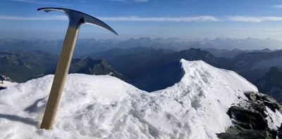An ice axe is stuck in the snow on a mountain peak overlooking a mountain landscape.