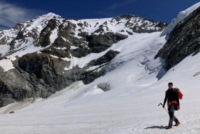 Person with ice pick and backpack on snow-covered mountain in front of rocky backdrop.