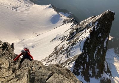 Person with helmet and climbing equipment climbs rocky mountain, snow-covered peaks in the background.