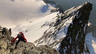 Person with helmet and climbing equipment climbs rocky mountain, snow-covered peaks in the background.