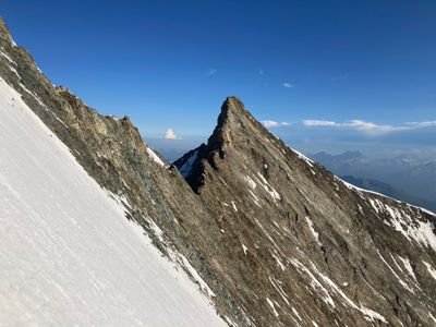 Mountain peak with a snow-covered slope and rocky summit, surrounded by blue sky and distant mountains.