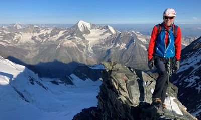 Person with helmet and climbing equipment standing on mountain peak, surrounded by snow-covered Alps.