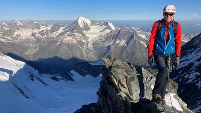 Person with helmet and climbing equipment standing on mountain peak, surrounded by snow-covered Alps.