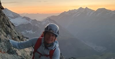 Person with helmet and climbing equipment on a rock wall, with mountains in the background during sunset.