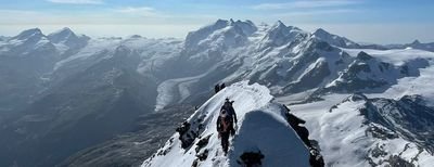 Mountaineers with helmets and ropes on a snow-covered ridge in the Alps, surrounded by mountains.