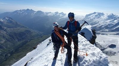 Two people with helmets and climbing gear are standing on a snow-covered mountain peak.