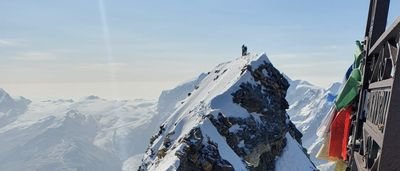 Mountaineer with helmet on snow-covered summit, surrounded by mountains; colorful flags in the foreground.