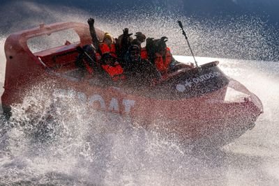 People in life jackets on a red speedboat, splashing through water, with mountains in the background.