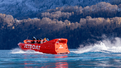 Red jet ski with people on a lake, surrounded by forested mountains and snow-capped peaks.