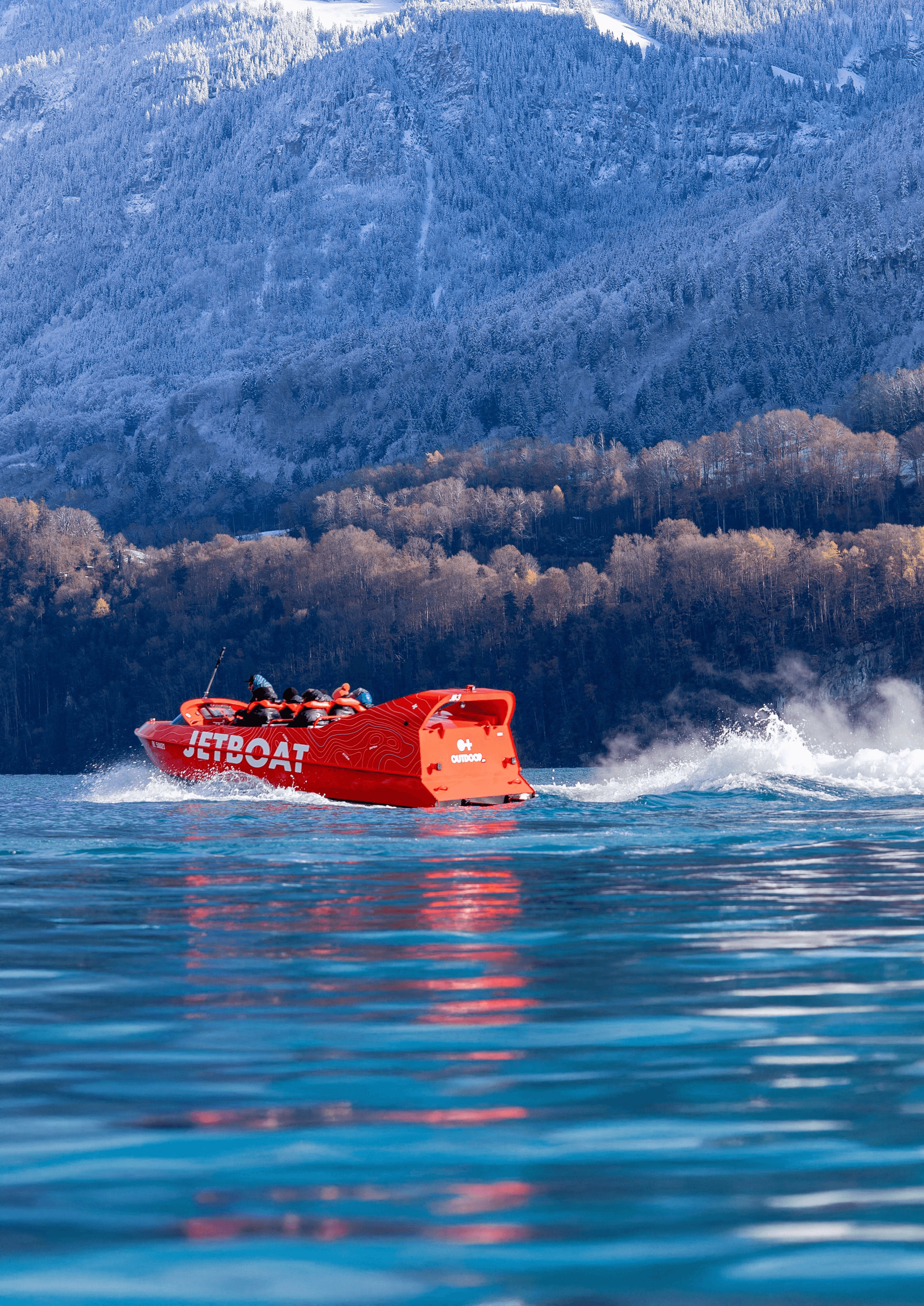 Red jet ski with people on a lake, surrounded by forested mountains and snow-capped peaks.