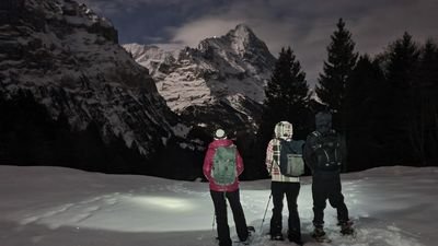 Three people with backpacks and hiking poles are standing in the snow in front of a mountain backdrop at night.
