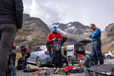 Group of climbers preparing gear and clothing near cars in a mountainous area with rocky peaks and snow patches.