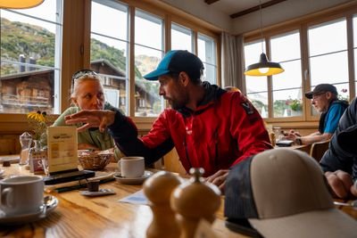 Two people in outdoor jackets having a discussion inside a wooden mountain cabin with large windows showing alpine scenery.