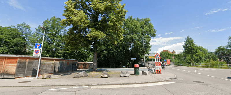 Empty street scene with road signs, trees, wooden fence, and a trash bin under a blue sky.