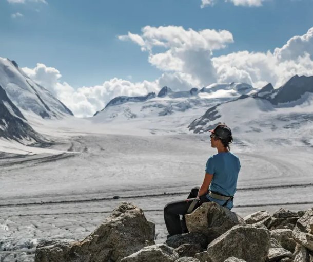 Person in helmet and harness sits on rocks, overlooking snowy mountains and glacier under cloudy sky.