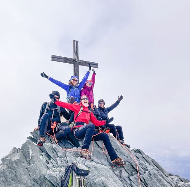 Five climbers in helmets and harnesses pose on a rocky mountain peak with a large cross.