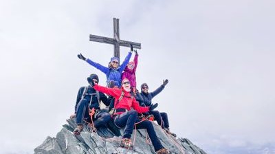 Five climbers in helmets and harnesses pose on a rocky mountain peak with a large cross.