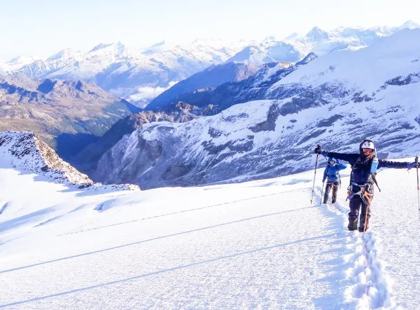 Two people in helmets and harnesses trek on a snowy mountain with poles, surrounded by peaks.