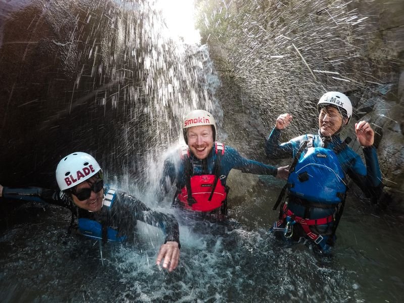 Drei Personen lachen unter einem Wasserfall beim Canyoning in einer Schlucht.