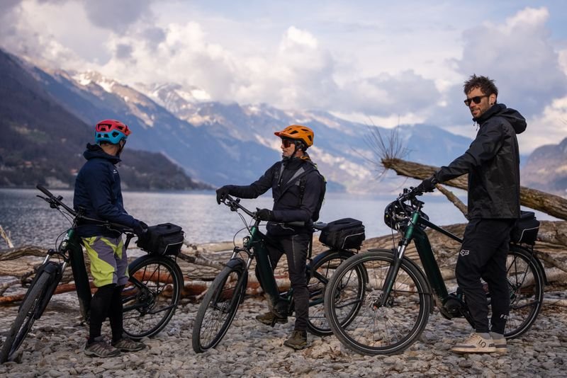Three people with helmets on e-bikes at the lakeshore, surrounded by mountains and clouds.