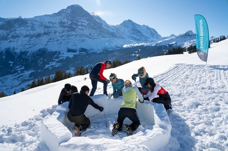 People are building an igloo in the snow, surrounded by mountains. Some are wearing winter clothing and hats.