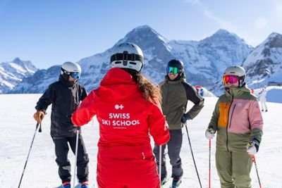 People with ski equipment and helmets are standing on a ski slope, surrounded by snow-covered mountains.