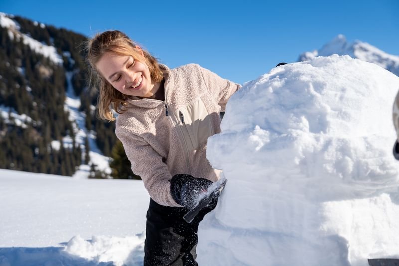 A person is building in the snow, wearing gloves; mountains in the background.