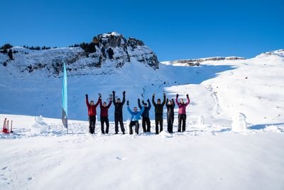 Personengruppe in Winterkleidung hebt die Arme auf schneebedecktem Berggelände vor Felswand.