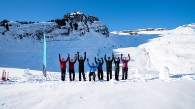 Personengruppe in Winterkleidung hebt die Arme auf schneebedecktem Berggelände vor Felswand.