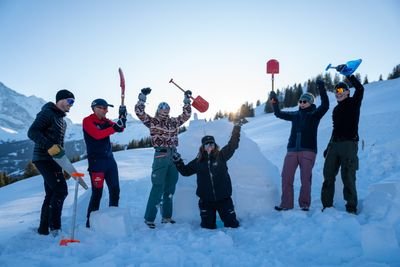 Six people with shovels in the snow, surrounded by mountains, wearing winter clothing and sunglasses.
