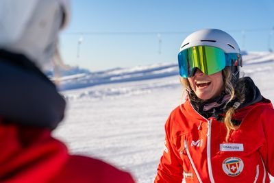 Personen in roten Jacken mit Helmen und Skibrillen stehen auf einer schneebedeckten Berglandschaft.