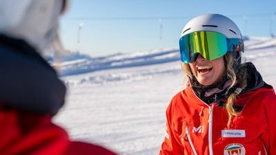 Personen in roten Jacken mit Helmen und Skibrillen stehen auf einer schneebedeckten Berglandschaft.
