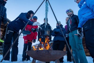Eine Gruppe von Menschen steht im Schnee um ein Lagerfeuer mit einem hängenden Kessel.